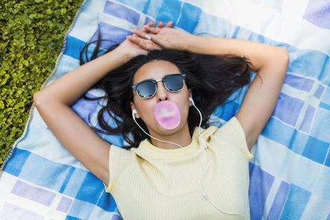 A young woman laying on a blue plaid blanket outside has sunglasses on, earbuds in, and is blowing bubblegum with her hands resting above her head.