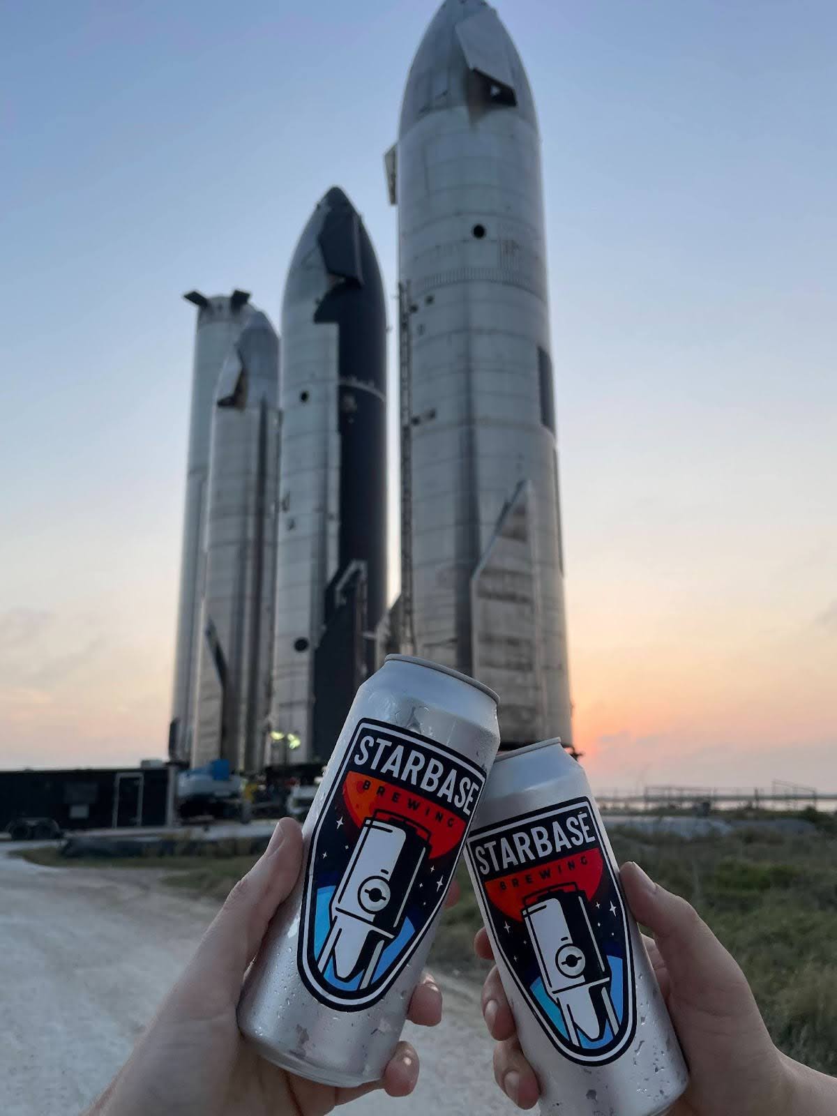 Two people cheersing with Starbase Brewing beers in the SpaceX Rocket Garden at sunset.