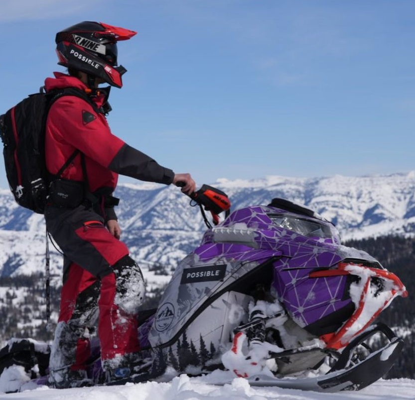 Person in red snow gear riding a purple snowmobile in a snowy mountainous landscape.