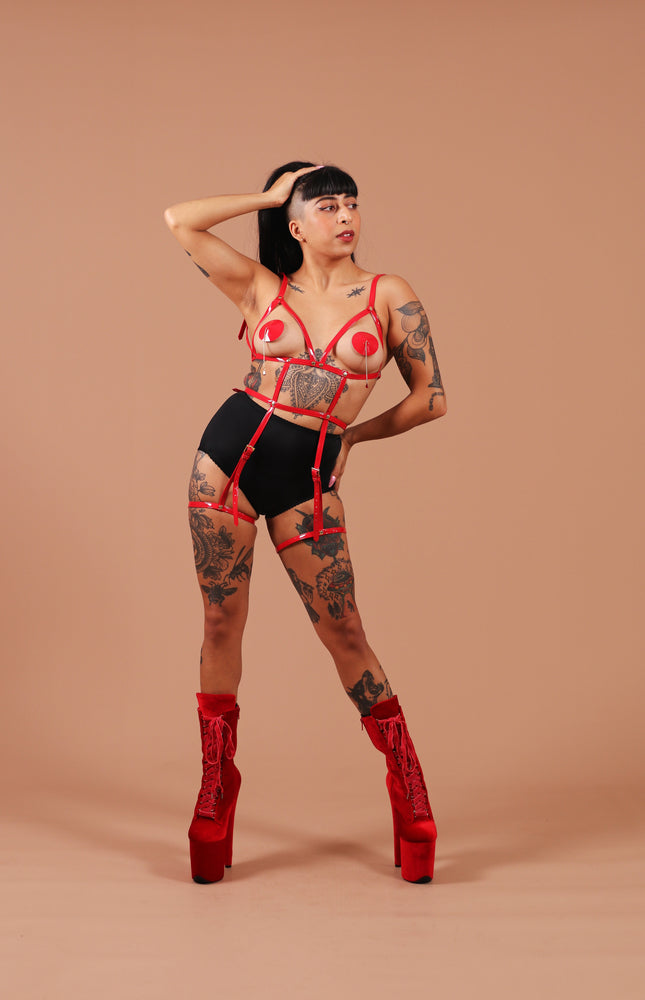 Mia is posing in the Red open harness bra with leg cuffs, black briefs and the bad blood red velvet boots. The harness cuts out around their breasts, framing them, which are complemented by red pasties. Two straps extend below the underbust of the open harness bra, connected to a waist strap that defines and cinches the waist, and further extending down to the thigh cuffs. The material is red and shiny, with small silver buckles on each of the straps.