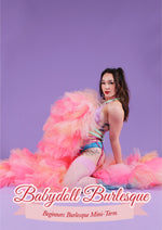 Babydoll Burlesque: Beginners burlesque mini-term. Tabby is kneeling on the floor against a purple background wearing pastel lingerie and pink stockings, covering her body with an oversized pink and coral tulle boa. She has long wavy brown hair in two pigtails, white skin and a slim frame.