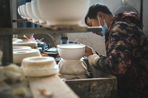 An experienced craftsman paints a ceramic bowl with steady hands, using a fine brush to carefully apply intricate details and patterns to the surface. The artist's skill and precision are evident in the meticulous strokes and attention to detail, creating a stunning work of art on the delicate ceramic surface.