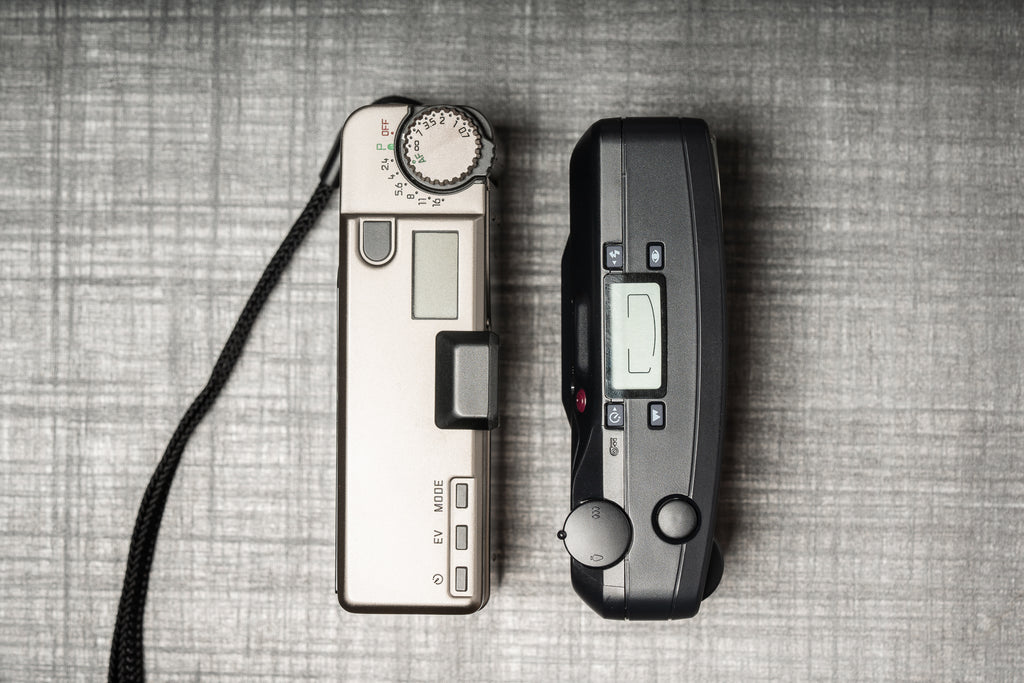 Leica Minilux to the left and a consumer film point and shoot to the right on a grey table