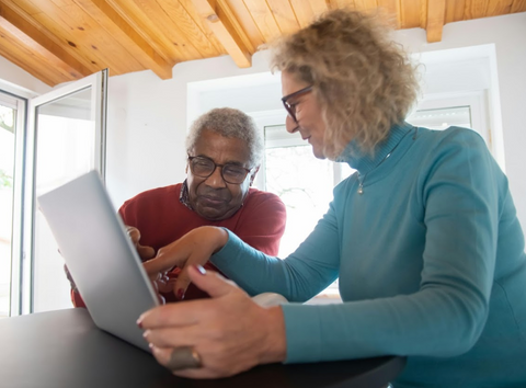 Elderly man and elderly women, both wearing glasses, look at laptop screen together and point.