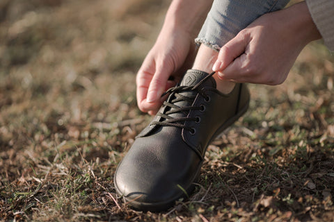 žena v barefoot botách Ahinsa shoes si na trávníku zavazuje tkaničky