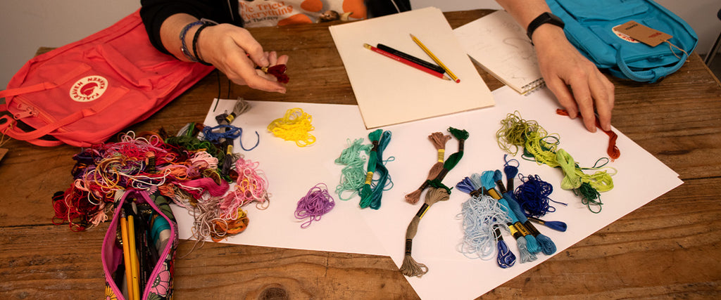 Craft table with colourful embroidery threads, Fjällräven backpacks, and sketching paper, during a Fjällräven customisation event.