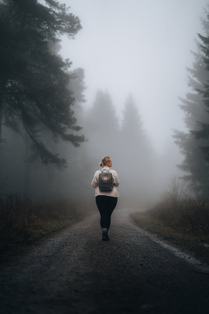 A solitary traveller walking down a misty forest road with a grey Fjällräven Kånken backpack, embodying the spirit of adventure in a serene natural landscape.