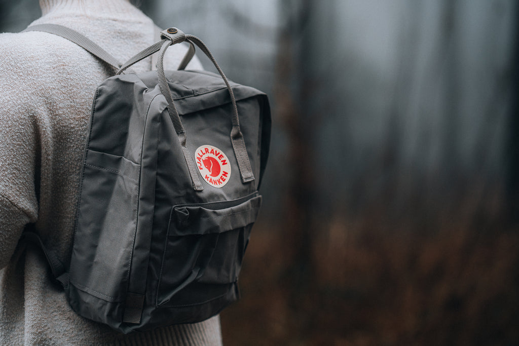Person wearing a grey Fjällräven Kånken backpack with a distinctive logo patch in a misty forest setting.