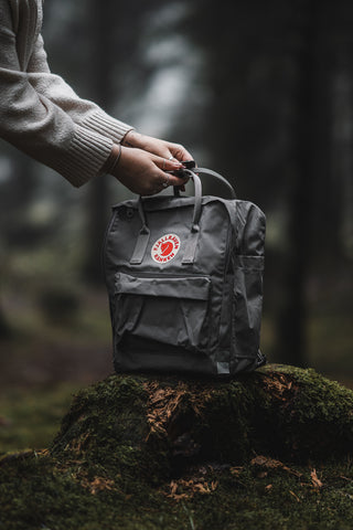 A hand lifting a classic grey Fjällräven Kånken backpack from a moss-covered stump in a dimly lit forest setting, showcasing the brand's iconic design and outdoor appeal.