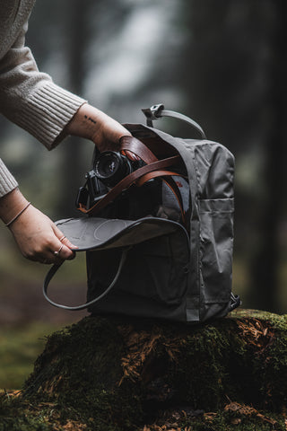 Close-up of a person’s hand opening a grey Fjällräven Kånken backpack to reveal a camera, set against the atmospheric backdrop of a dense forest.