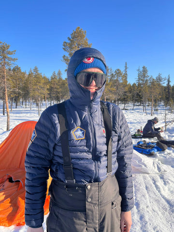 Outdoor enthusiast Tom Jacques-Milner in a Fjällräven down jacket with hood and branded patches, ready for cold weather, with a snowy campsite in the background.