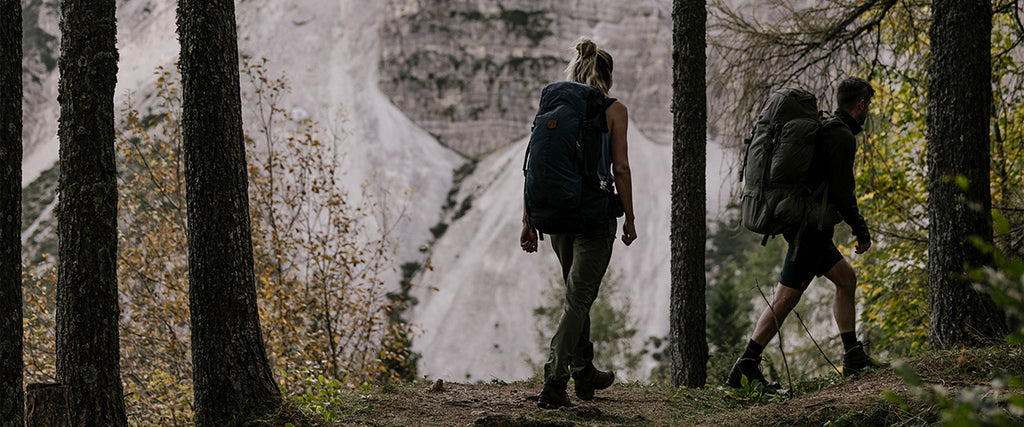 Two hikers trekking through a forest trail with Fjällräven backpacks, the brand's logo clearly visible on their gear, set against a backdrop of a dramatic mountainous landscape.