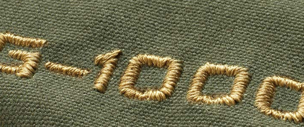 Close-up of the golden embroidered Fjällräven G-1000 logo on a green fabric, highlighting the quality and detail of the brand's signature stitching.