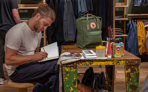 Artist sketching designs in a Fjällräven store with a custom-painted Kånken backpack and painting supplies on the table