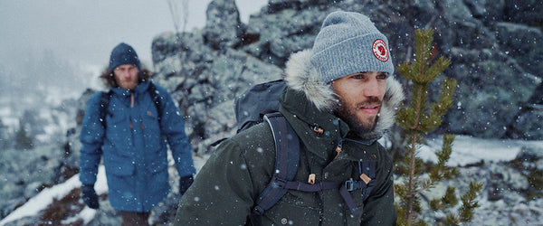 Two hikers in a snow-covered landscape, forefront man wearing a Fjällräven green parka with fur-lined hood and a beanie, and companion in a blue Fjällräven jacket.