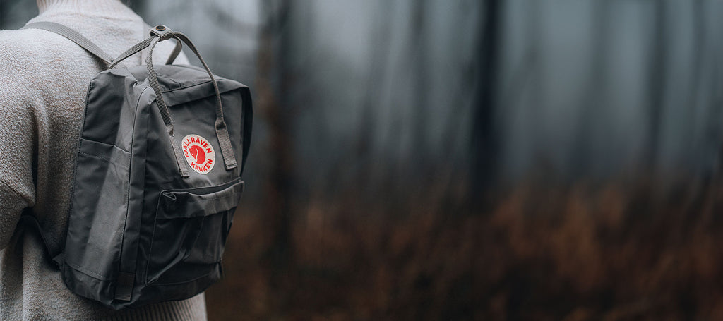 Close-up of a classic grey Fjällräven Kånken backpack worn by a person against a blurred forest backdrop, focusing on the iconic red and white logo patch.