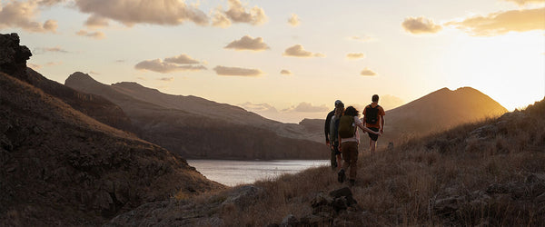 Three hikers with Fjällräven gear trekking at sunset through rugged terrain with a view of a serene lake in the distance, highlighting the adventurous spirit of the brand.