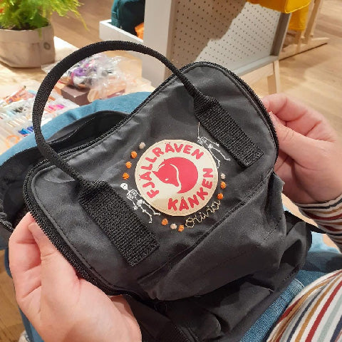 Hands presenting a black Fjällräven Kånken mini backpack with custom embroidery around the classic logo.
