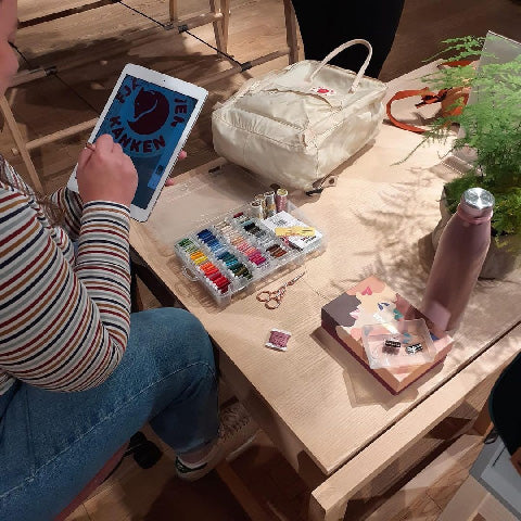 Person embroidering a custom design on a Fjällräven Kånken backpack, with embroidery threads and tools on a wooden table.