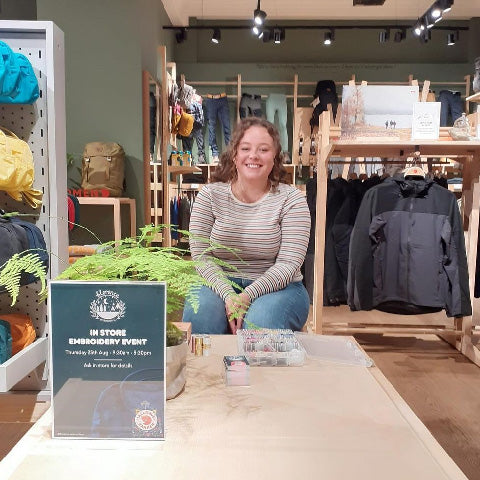 Smiling woman sitting at a Fjällräven store during a custom embroidery event, with a sign and embroidery materials on the table.