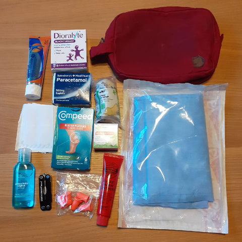 Essential first-aid supplies for hiking, including Dioralyte sachets, paracetamol, blister plasters, antiseptic cream, bandages, a red Fjällräven bag, and sanitation items neatly displayed on a wooden surface.