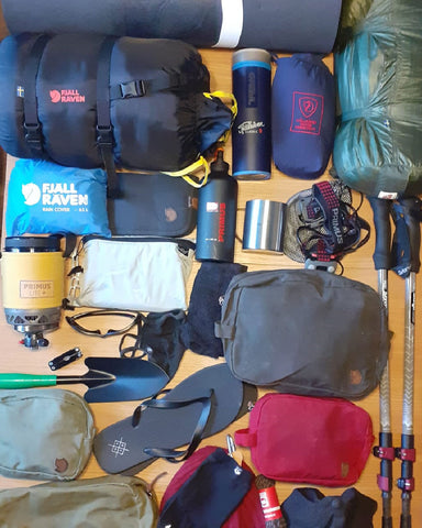 A variety of hiking equipment laid out, including Fjällräven sleeping bags, backpacks, a Primus stove, trekking poles, and thermal flasks, prepared for an outdoor adventure.