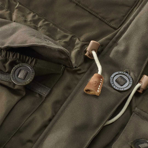 Close-up of a Fjällräven 'Numbers' jacket detail, showing the leather zipper pull and the brand's signature black and grey Arctic fox logo patch on olive green fabric.