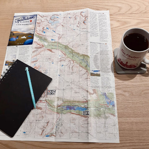 Planning a hike with a Fjällräven map, notebook, pen, and a mug of tea on a wooden table, suggesting preparation for an outdoor trek.