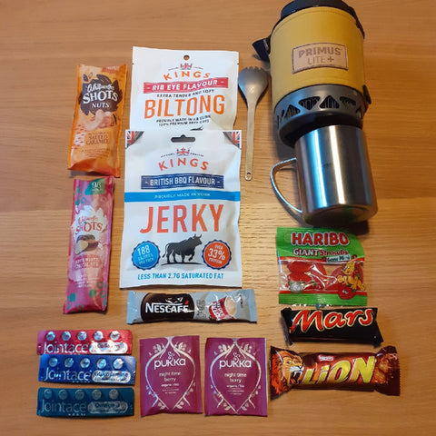 Assorted hiking snacks and drinks including nuts, biltong, jerky, energy bars, candy, instant coffee, tea bags, and a Primus stove mug, arranged on a table.