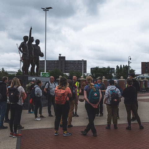 Group of hikers with Fjällräven backpacks gathered at Manchester United's Old Trafford, with the famous United Trinity statue prominent in the background.