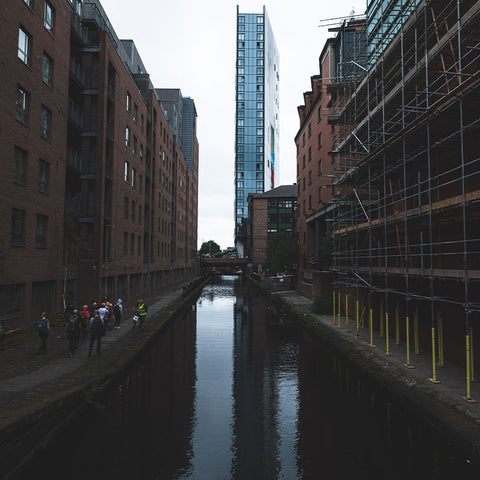 A Manchester Fjällräven Walk with people strolling by a canal lined with modern and traditional architecture under construction, reflecting urban development and outdoor activity.
