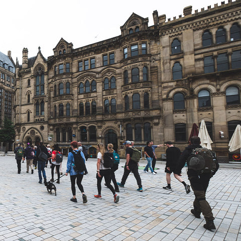 People walking across a square in Manchester in front of a historic building, one individual prominently carrying a black Fjällräven backpack