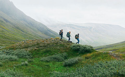 Three hikers with Fjällräven backpacks trekking through a mountainous landscape with lush greenery.