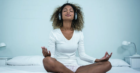 Female on bed in white walled room meditating legs crossed hands with hands relaxed and headphones on listening to musi