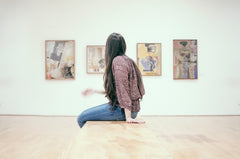 a woman with long brown hair is sitting on a bench looking at artwork on a gallery wall behind her