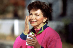 older woman in a pink top is outdoors holding a bottle of sun screen. She has her other hand up to her face putting sunscreen on 
