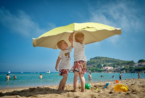 Two young boys in swim suits and rash guards are setting up an umbrella on a beach