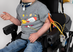 closeup of boy seated in wheelchair wearing spaceship g-tube zip shirt opened with feeding tubes coiled with cath clip