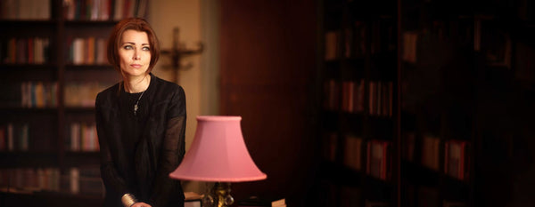 A photo of author Elif Shafak, showing her sat behind a pink lamp in a tastefully furnished room with a large bookshelf.