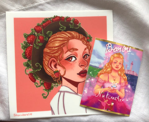 Photo of two stickers, one being a redraw of the cover of Barbie Nutcracker and the other, bigger one, being a white woman with strawberry blonde hair pulled into a hat with strawberries growing on top.