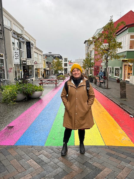 Tuija wearing a brown raincoat stands in front of a rainbow painted street in Reykjavik.