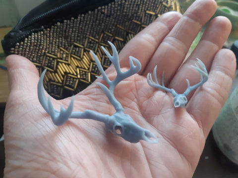 3 tiny 3d-printed white-tailed deer skulls resting in the palm of someone's hand. One is 20% larger than the other two because they will be made into a pair of earrings and a necklace to be painted in neo woodland style and coated in resin.