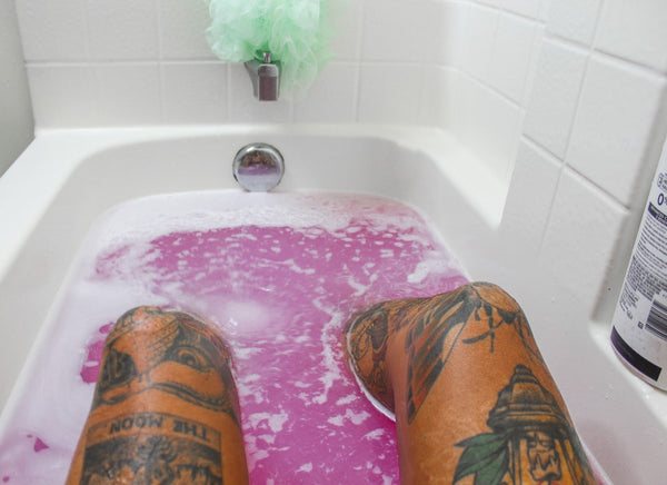 A person with tattoos on their legs relaxes in a bath tub with purple water from a bath bomb