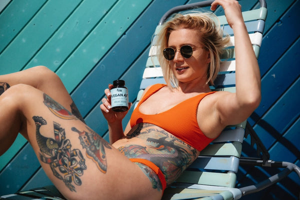 A girl with tattoos and sunglasses lays hungover by the pool on her lounge chair holding a bottle of Sunday Scaries Vegan AF gummies