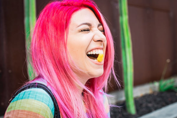 A woman with pink hair and a gummy in her mouth laughs and smiles after hearing a joke