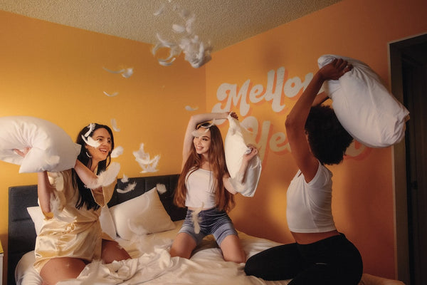Three girls have a pillow fight on a bed in a yellow room