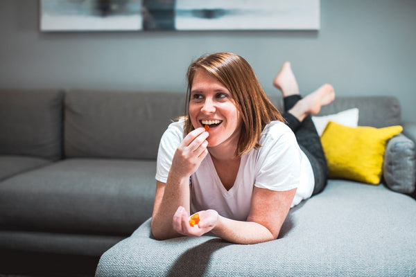 A girl lies down on her sectional couch while watching a movie and eating a Sunday Scaries gummy