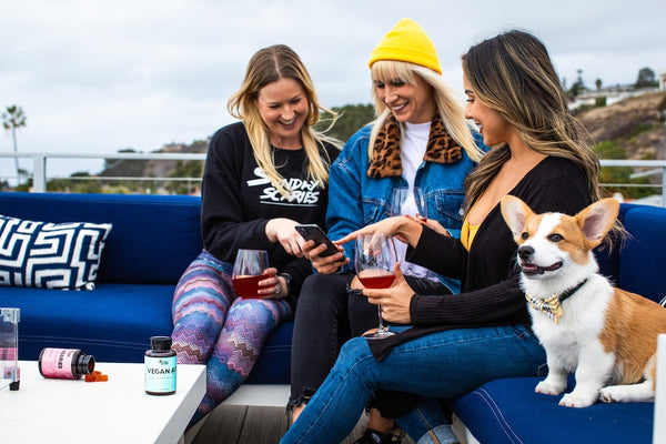 Three girlfriends sit on a rooftop and share something on their phone while sitting with their dog and drinking wine