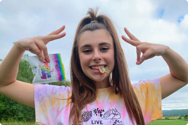 A happy girl holds up two peace signs with her hands while a piece of Sunday Scaries CBD Candy, called Unicorn Jerky, hangs from her mouth