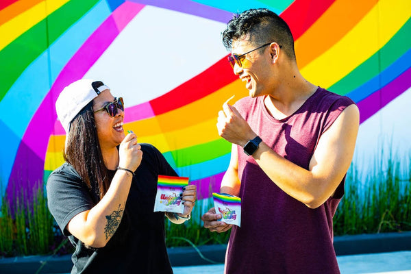 A woman and man embrace pride by eating Unicorn Jerky in front of a rainbow wall